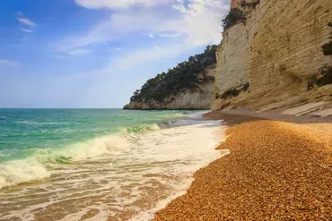 Spiaggia di ciottoli e mare con scogliera bianca e cielo blu.