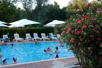 Piscina con giocatori, sedie a sdraio e ombrelloni bianchi. Fiori rossi in primo piano.