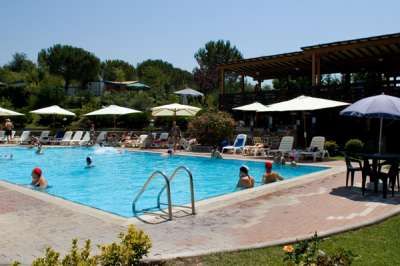 Piscina con persone, lettini e ombrelloni. Edificio sullo sfondo.