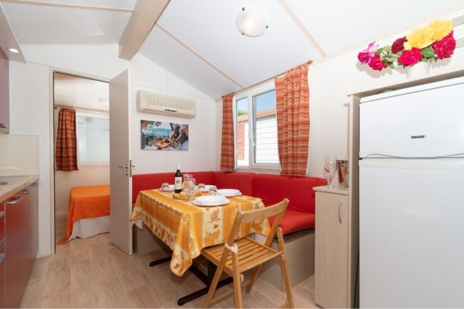 Interno, tavolo con tovaglia gialla, sedie, frigorifero e divano rosso.