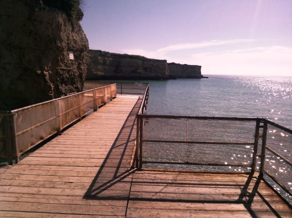 Passerella in legno su una costa rocciosa che si affaccia sul mare calmo e brillante.
