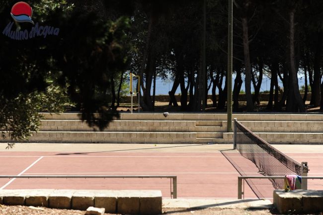 Campo da tennis all'aperto, circondato da alberi e panchine.