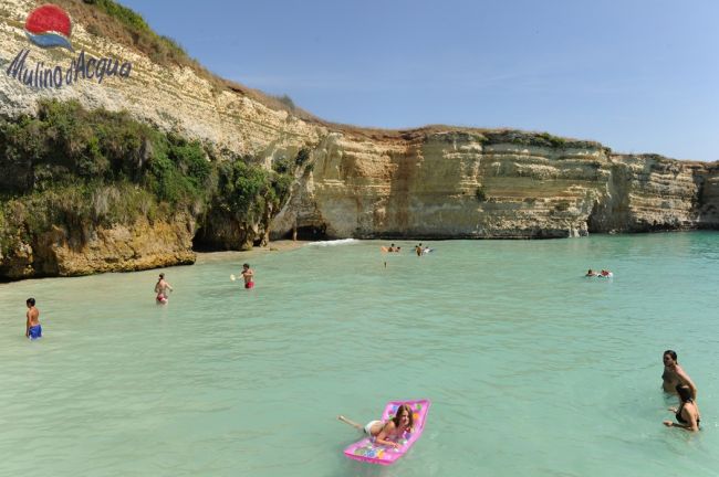 Persone nuotano in un mare turchese vicino a scogliere rocciose.