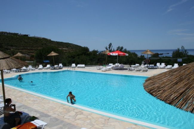 Piscina all'aperto con ombrelloni di paglia e lettini. Vista collinare e mare sullo sfondo.