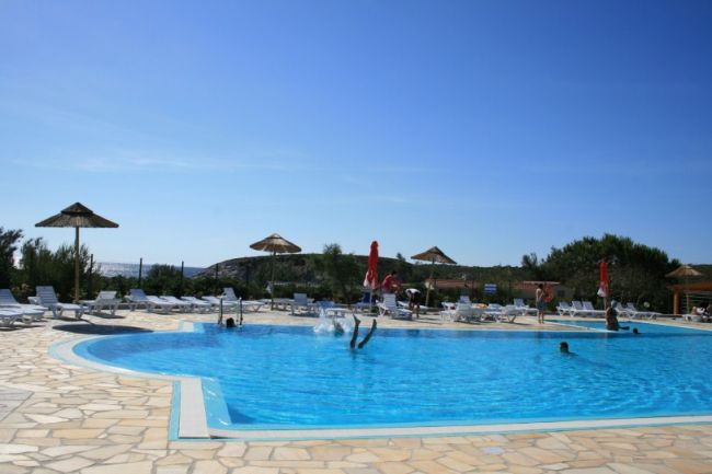 Persone che nuotano in una piscina con lettini, ombrelloni di paglia e cielo sereno.
