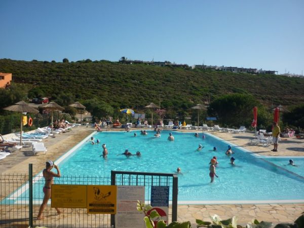 Piscina affollata con bagnanti, lettini e ombrelloni di paglia, colline verdi sullo sfondo.