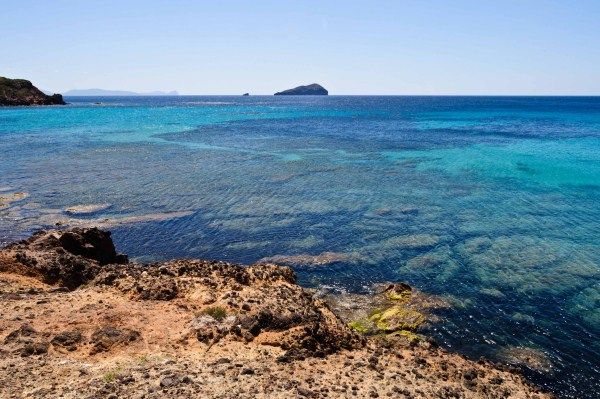 Scogliera rocciosa con mare cristallino, tonalità blu e verde, isola in lontananza.