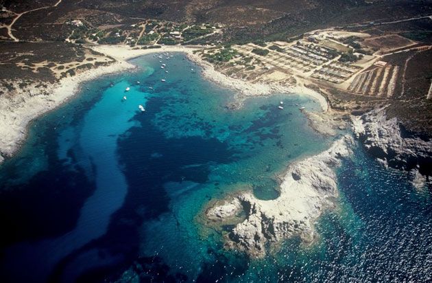 Veduta aerea di una costa con acque blu, barche e terreno arido circostante.