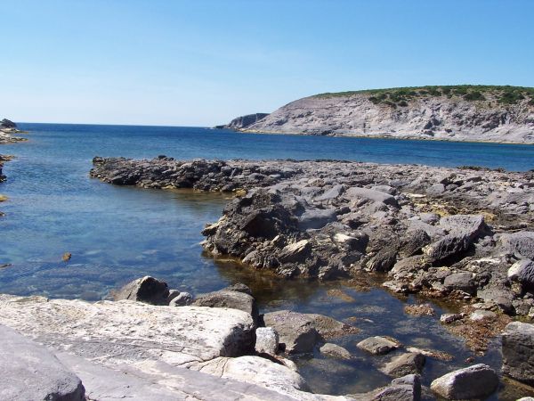 Paesaggio costiero roccioso con acqua limpida, promontorio in lontananza.