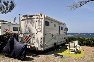 Un camper parcheggiato vicino al mare con sedie a sdraio e tavolino su un tappeto verde.