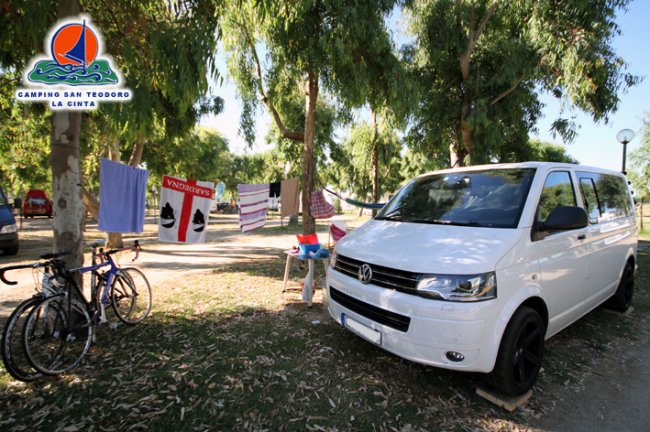 Un furgone bianco vicino a biciclette e panni stesi tra gli alberi.
