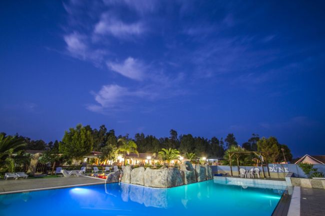 Piscina illuminata di sera, circondata da piante e un cielo blu scuro.