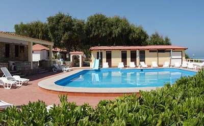 Piscina all'aperto circondata da lettini, vegetazione e case vacanza.