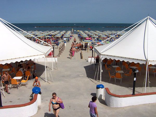 Spiaggia con file di ombrelloni e sdraio, tra due tendoni bianchi con tavoli.