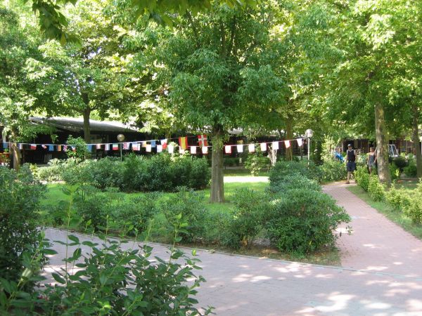Giardino verde con siepi, alberi e stradina di mattoni. Bandiere colorate appese tra gli alberi.