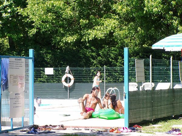 Persone si rilassano e giocano in piscina sotto gli alberi.
