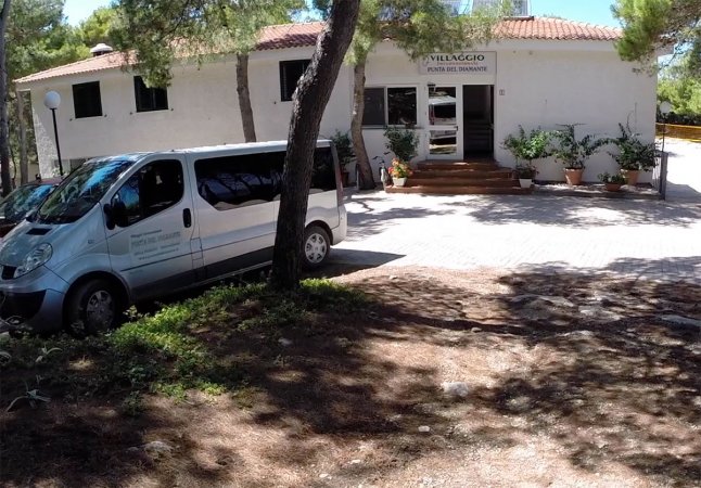 Edificio bianco con un furgone parcheggiato davanti, circondato da alberi.