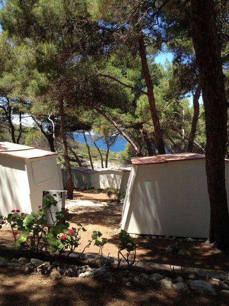Campeggio tra alberi con vista sul mare, piccole strutture bianche e fiori.