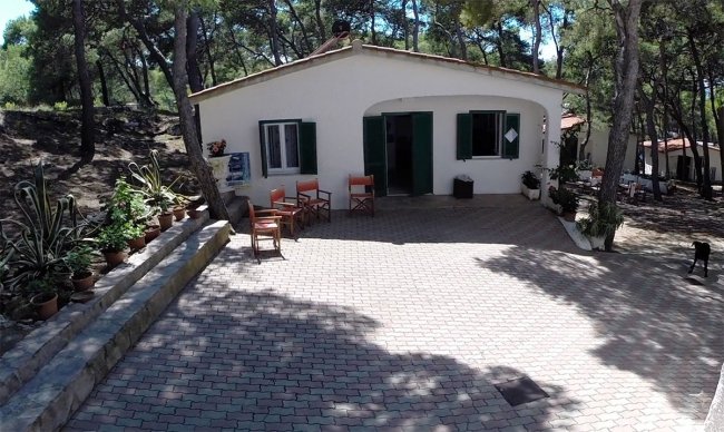 Casa bianca con patio pavimentato, sedie rosse, piante e alberi intorno.