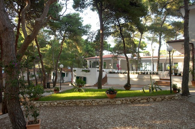 Un edificio bianco tra alberi, con giardino in primo piano e scale che portano a una terrazza.