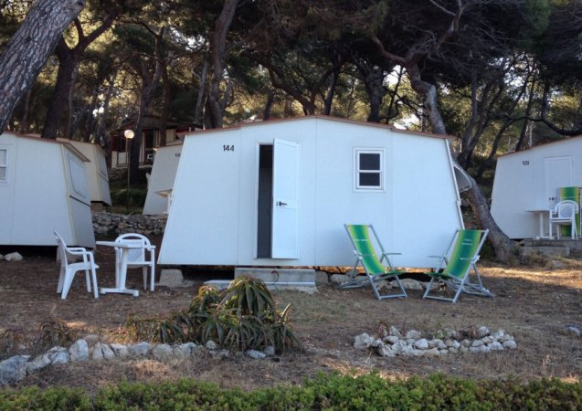 Casetta bianca con sedie a sdraio verdi e tavolo, immersa tra gli alberi.