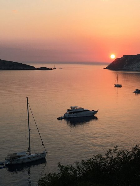 Tramonto su una baia con barche e yacht su acque calme, cielo arancione e sole vicino all'orizzonte.