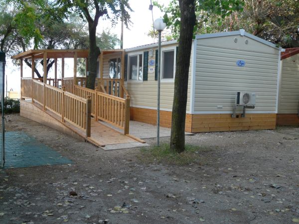 Casa mobile bianca con veranda in legno circondata da alberi.