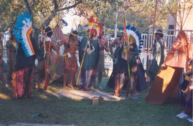 Persone in costumi tribali colorati radunate in cerchio su un prato con alberi.