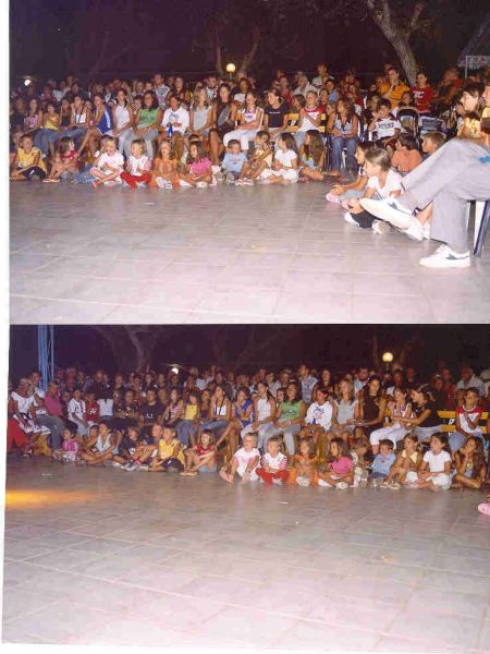 Un pubblico seduto all'aperto, composto da adulti e bambini, osserva qualcosa fuori campo.