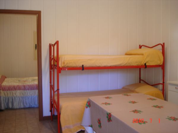 Letto a castello rosso con coperte gialle accanto a un tavolo con tovaglia a fiori, pareti bianche.