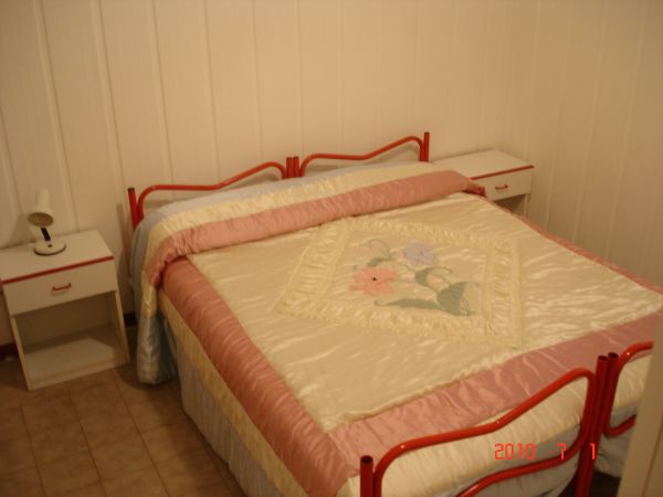 Letto matrimoniale con copriletto rosa e bianco, due comodini bianchi con lampade.