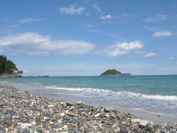 Spiaggia di ciottoli con mare blu e un'isola in lontananza sotto un cielo parzialmente nuvoloso.