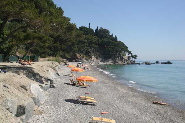Una spiaggia di ciottoli con ombrelloni arancioni e vista sul mare. Alberi e rocce sullo sfondo.