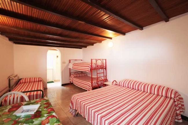 Camera con letto matrimoniale, letti a castello e arredi a righe rosse. Tavolino e sedie.