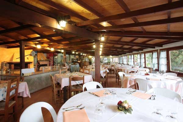 Sala ristorante con tavoli rotondi apparecchiati, grandi finestre e soffitto in legno.