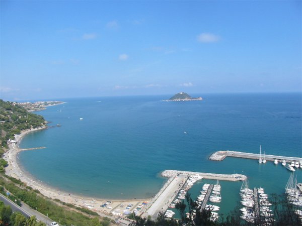 Vista di una baia con mare blu e un'isoletta. Un porto turistico è visibile sulla destra.