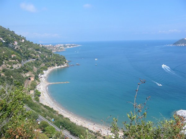 Spiaggia curva con mare blu e colline verdi. Una barca crea scie bianche nel mare.