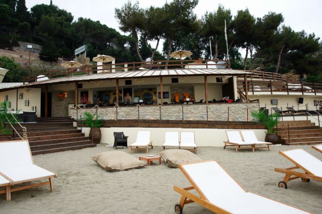 Spiaggia con lettini e bar in legno sullo sfondo.