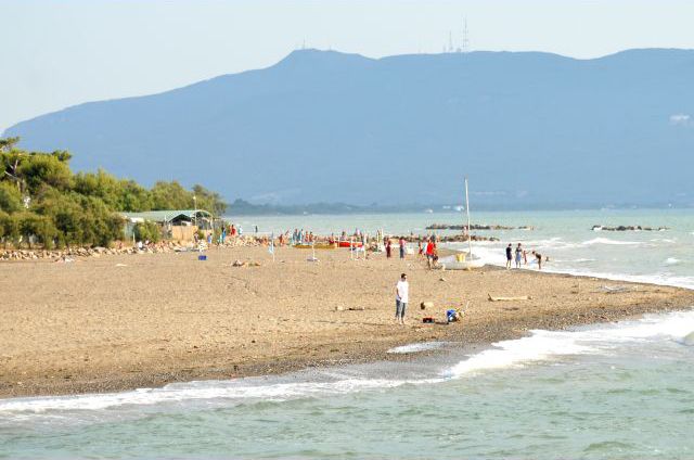 Spiaggia sabbiosa con bagnanti, alberi e colline sullo sfondo.