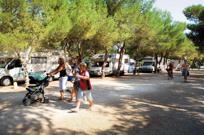 Persone passeggiano tra camper in un'area alberata.