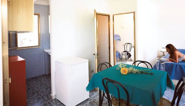 Una sala con tavolo coperto e sedie, piccola cucina visibile.