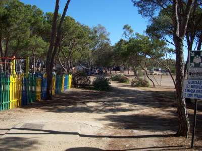 Strada sterrata tra alberi, con un parco giochi a sinistra e un parcheggio in lontananza.