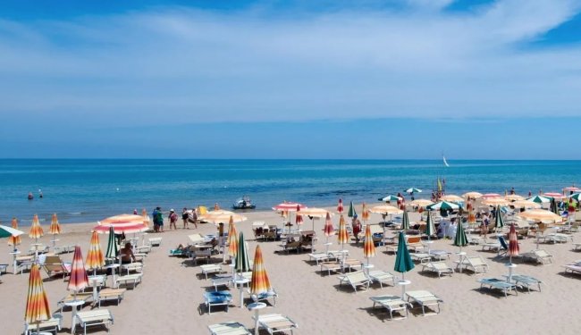 Spiaggia sabbiosa con ombrelloni colorati e sdraio, vista mare blu e cielo sereno.