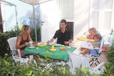 Una famiglia di quattro persone fa colazione su una terrazza, seduti attorno a un tavolo con cibo.