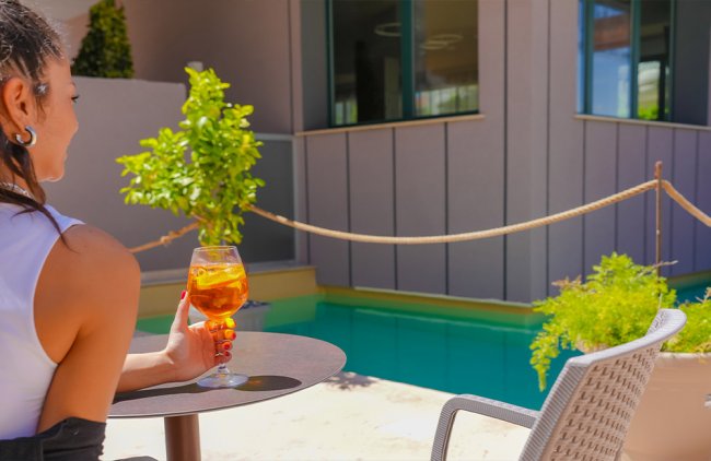 Persona seduta all'aperto, tiene un drink vicino a una piscina e piante verdi.