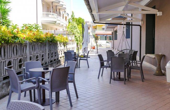 Spazio esterno con tavolini, sedie e ombrelloni, circondato da piante.