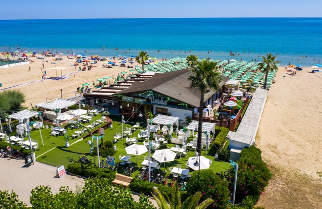 Vista aerea di una spiaggia con ombrelloni verdi e un lido con giardino e palme vicino al mare.