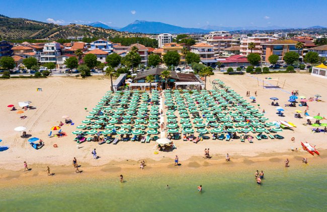 Spiaggia affollata con file di ombrelloni verdi, mare e case in lontananza.