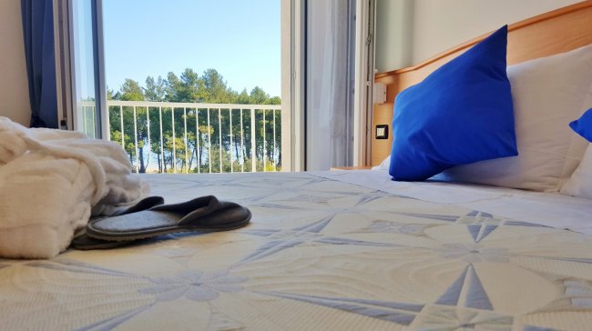 Camera da letto vista mare, cuscini blu su letto, accappatoio e ciabatte in primo piano.