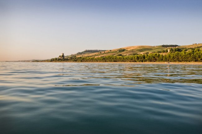 Mare calmo con costa verdeggiante e colline lontane, cielo limpido.